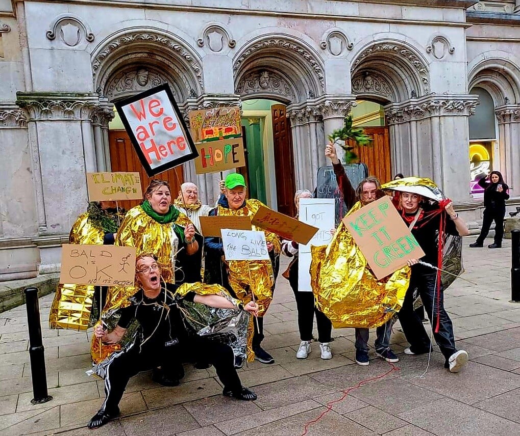 members of The Black Box wearing gold foil and holding signs including one saying "we are here"